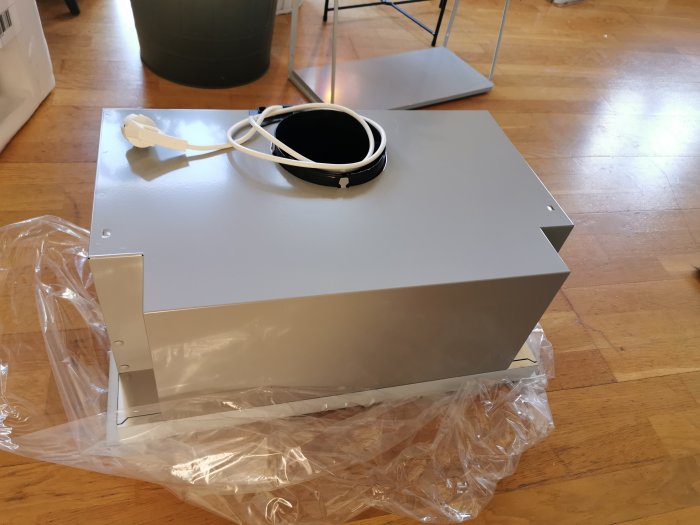 En ny köksfläkt urpackad på ett trägolv, med plastemballage och ett elkabel.