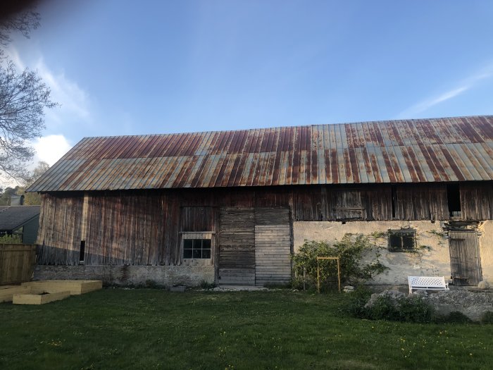 Äldre ladugård med slitet och rostigt plåttak i olika nyanser av rött och brun med frågetecken kring dess hållbarhet.