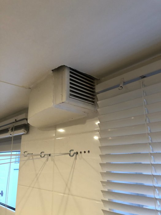 Tilluftventil i hörn av ett oisolerat uterum ovanför vita persienner.