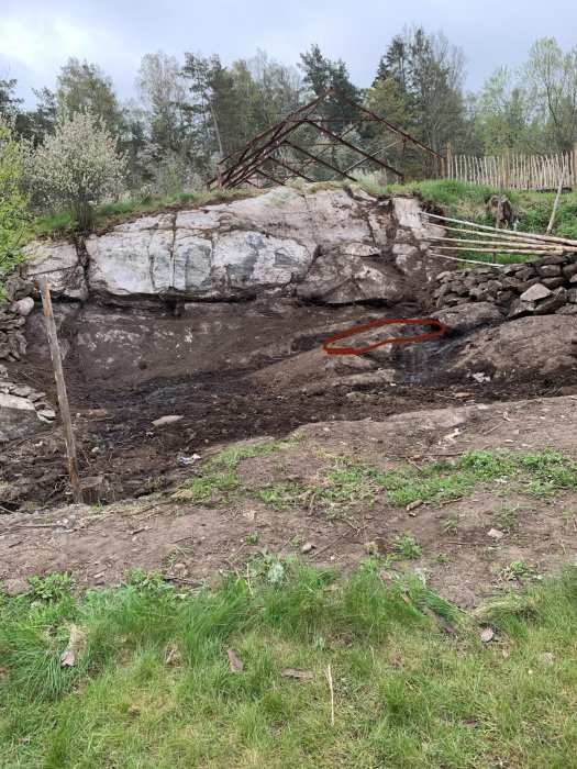 Grävd yta upp till berget med röd ring markerande högsta punkten, omgivet av naturlig vegetation och en rostig växthusstomme.