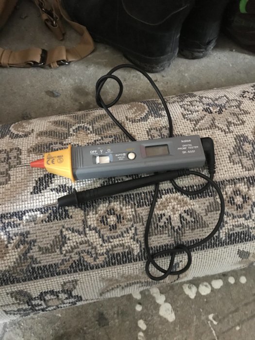 Digital multimeter ligger på ett mönstrat tyg nära ett par slitna arbetsstövlar.