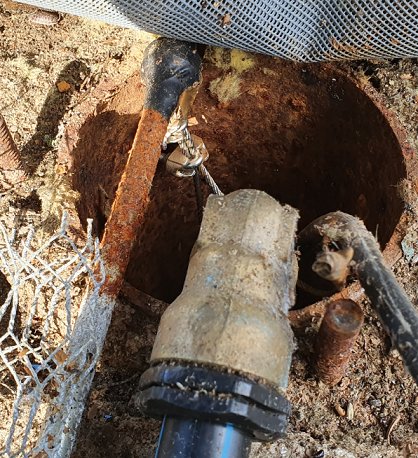 Rostiga rör och kablar i ett 120mm borrhål för dricksvatten, omgivet av jord och nät.