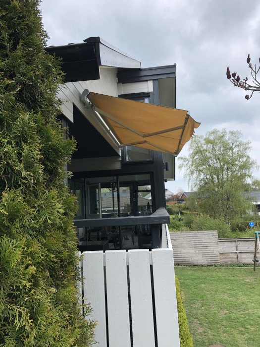 Uppskruvad markis med guldfärgat tyg på ett modernt husfasad, omgiven av grönska och staket.