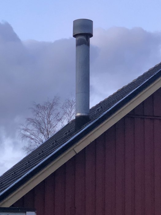 En metallskorsten som sticker upp från ett rött hus med svarta takpannor i skymningen.