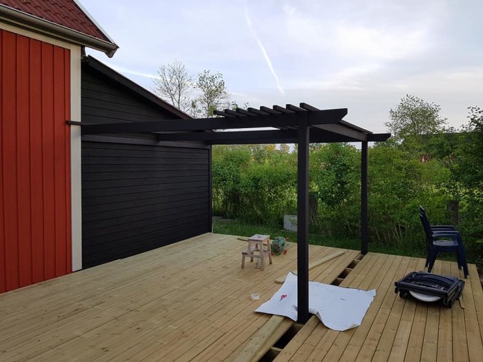 Trädäck med pergola vid en röd och svart husfasad, trädgårdsmöbler och grönska i bakgrunden.