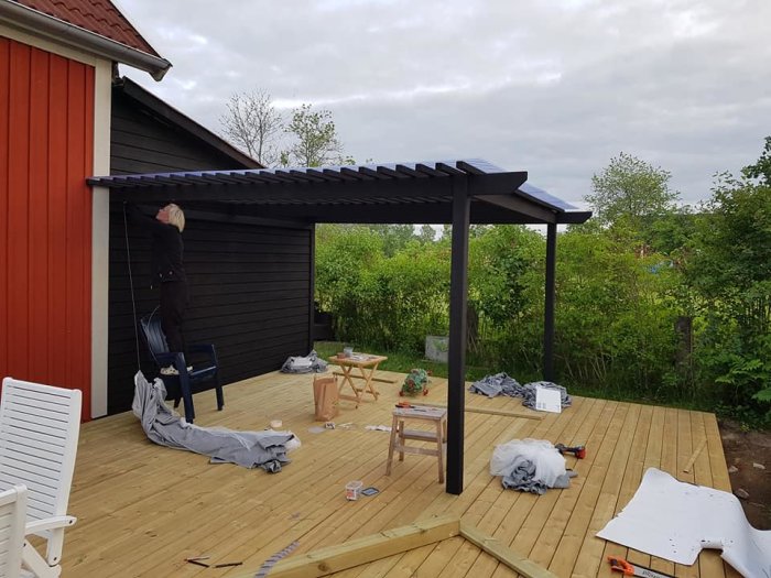 Person som bygger en träaltan med svart pergola nära röd husvägg, verktyg och byggmaterial syns.