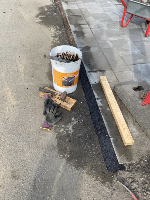 Reparerat asfaltsområde med reparationsasfalt, hink, handskar och verktyg på en uppfart.