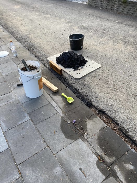 Reparationsarbete på uppfart med asfalt, hink, spade och snittmärkning på trottoarsten.