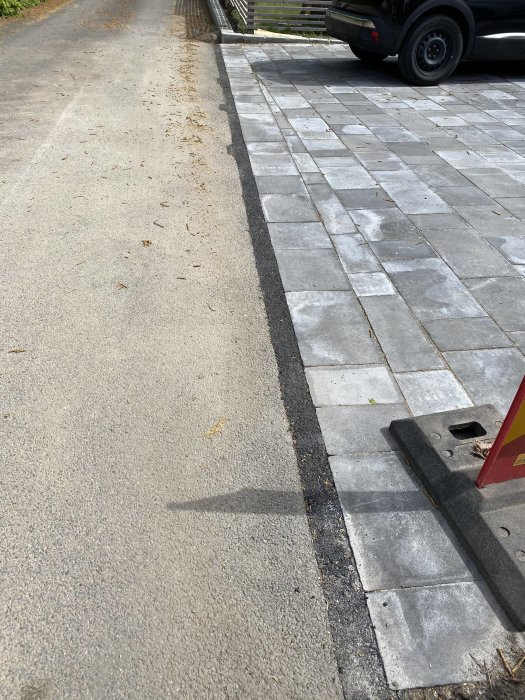 Asfaltsyta nära en rad stenplattor, med en tydlig rak skarvningslinje, och en parkerad bil delvis synlig.