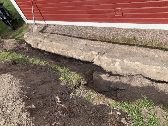 Fallen betongstödmur längs med husvägg, omgiven av jord och gräs.
