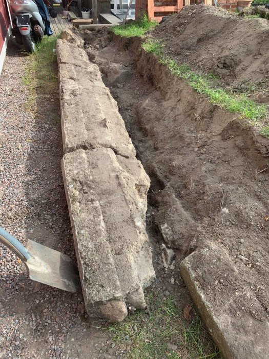 Fallen stödmur av betongblock vid en grävd jordsänka, intill skottkärra och landskap.
