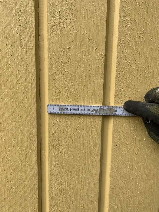 En hand håller ett måttband mot en gul huspanel för att mäta tjockleken, troligtvis en äldre typ av fasadpanel.