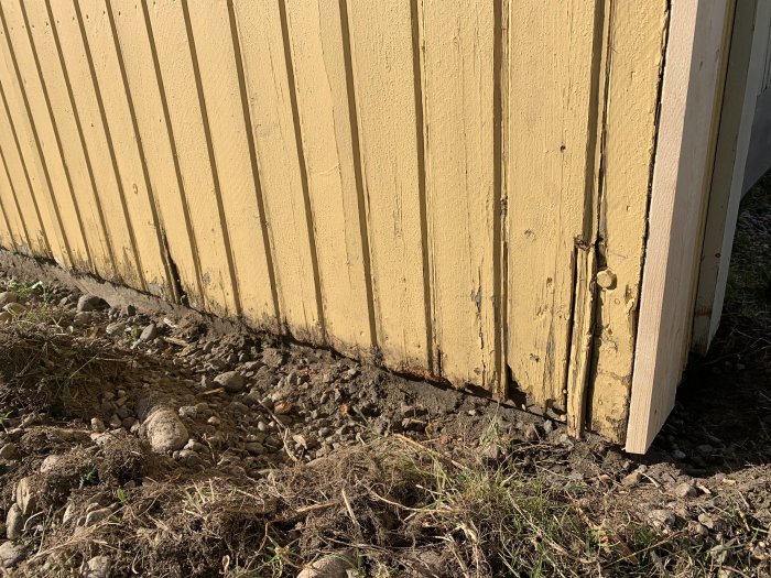 Skadad gavel på en gul träfasad med sprickor och fuktskador vid nedre kant över ojämn mark med småstenar.