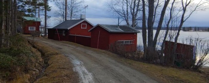 Lantligt rött hus och uthus nära en frusen sjö synliga bakom träd, med en grusväg i förgrunden.