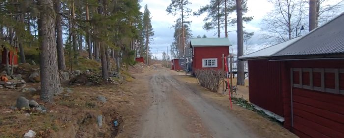 Grusväg passerar röda trähus och uthus med skogsbakgrund som avgränsar gammal åkermark.