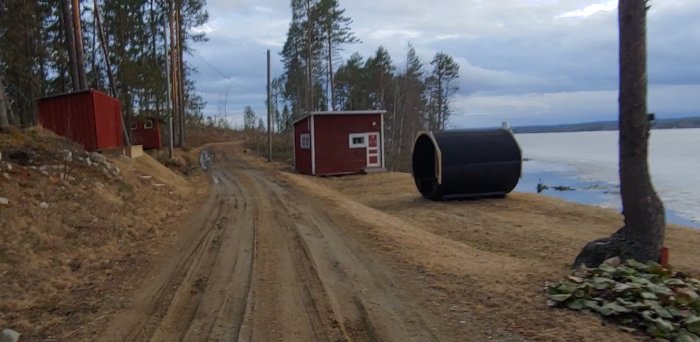 Landsväg nära vatten med röda uthus och svart tunnformad konstruktion, gräs och stenar i förgrunden.