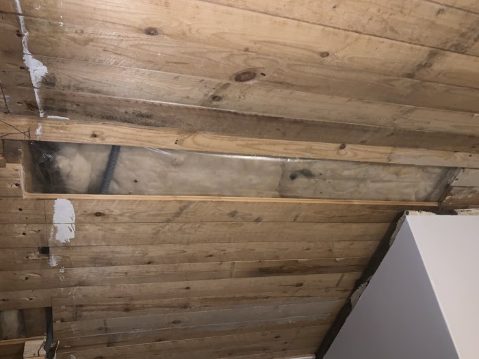 Öppnat innertak i ett gammalt hus visar träbjälkar och svart isolering med en synlig reva i plasten.