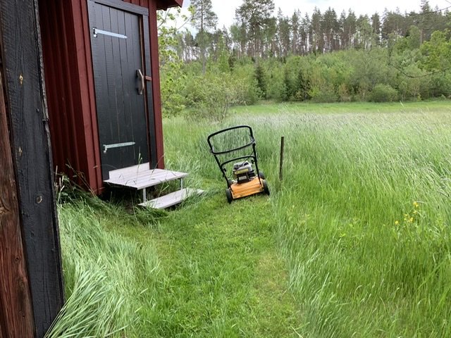 Friliggande litet rött uthus med högt tak vid en äng, omgivet av högt gräs med en åkgräsklippare framför.