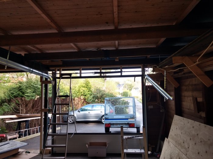 Inredning av ett garage under konstruktion med träreglar och bärlina, synlig I-balk och material på golvet, utsikt mot en parkerad bil.