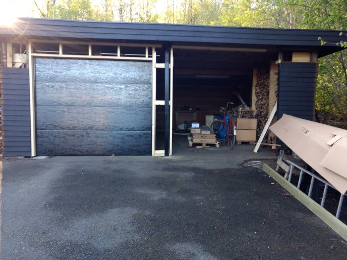 Halvfärdigt garage med öppet tak, delvis stängd port och diverse föremål och verktyg inuti.