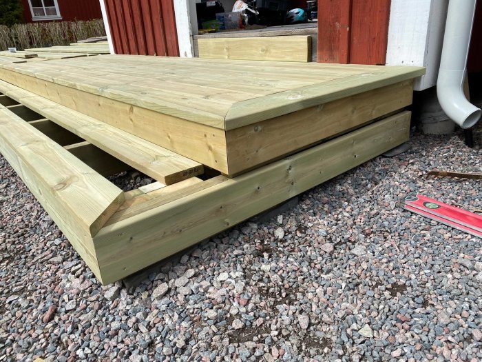 Trätrall med ett mellanrum mot en byggnad och en vattenpass på grus nära regelverket.