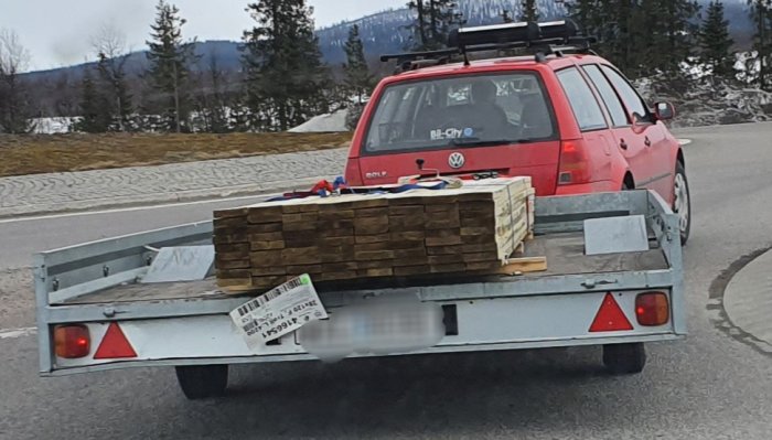 Röd bil med släpvagn lastad med löst timmer som kan vara farligt vid inbromsning.