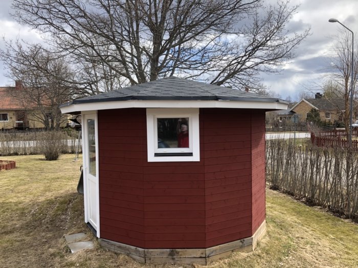 Rödmålad lekstuga med svart tak, likt ett lusthus på ena sidan, står i trädgård.