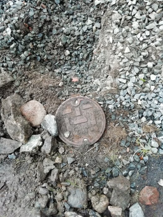 Grävd mark med ett övervuxet brunnslock och dräneringsrör, omgivet av makadam och jord, nära en husfasad.
