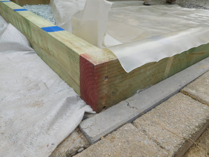 Närbild av nyligen gjuten betongplatta med ohyvlat impregnerat trä som gjutram och vit plastfilm ovanpå.