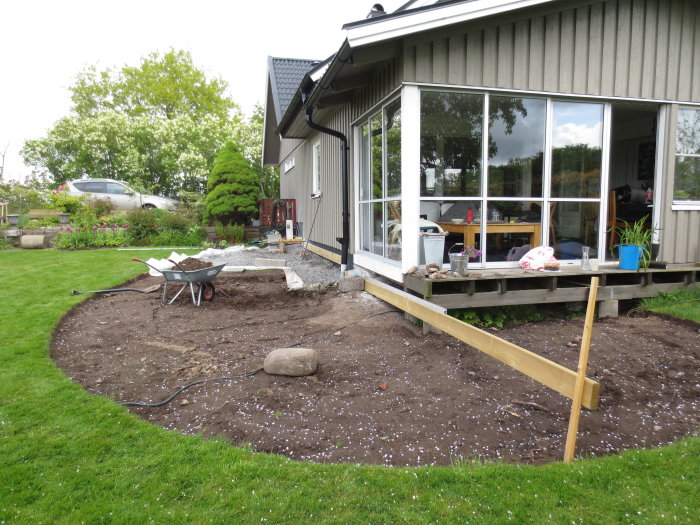 Trädgårdsbygge med en nylagd träram för trädäck intill husets veranda, en skottkärra, och blommande trädgård i bakgrunden.