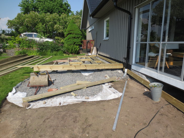 Byggplats vid hus med början till trädäck, med träreglar, grus och verktyg utspridda på marken.