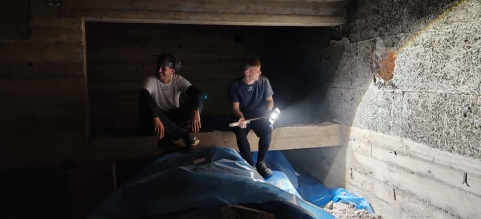 Två personer undersöker ett nyupptäckt utrymme bakom en vägg i en källare med bara ficklampor som ljuskälla.