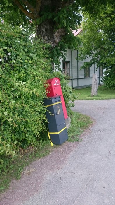 Två lutande brevlådor intill varandra vid en vägkant, en i rött och svart, den andra i svart och gult.
