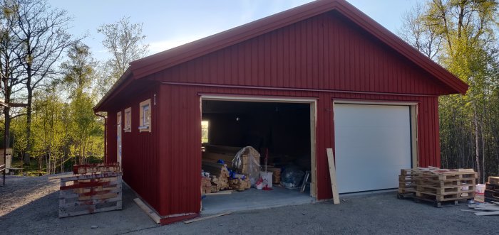 Röd trädgårdsbod med en öppen och en stängd vit garageport, byggmaterial synligt inuti.