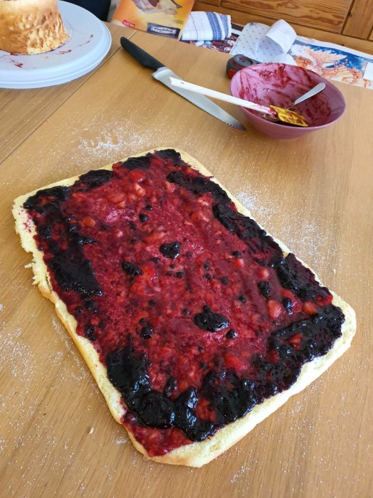 Tårta under tillverkning med jordgubbar och sylt på ett köksbord, bakredskap i bakgrunden.