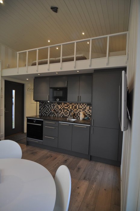 Modernt kök med mörka skåp och loft med vita räcken ovanför, betonar effektivt utnyttjande av utrymme.