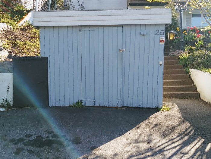 Lutande gammalt cykelförråd med sliten blå färg på asfaltsunderlag bredvid betongtrappa.