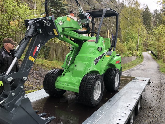 Grön Avant 423 minilastare på en släpvagn bredvid en man, i ett villaområde.