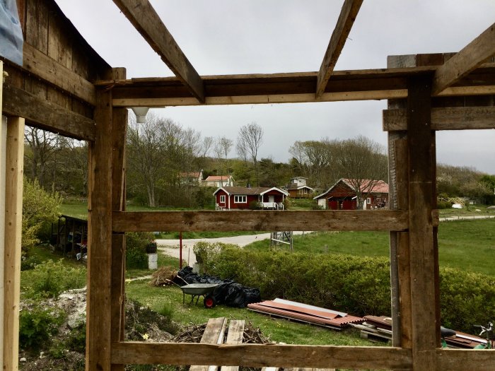 Utsikt genom en demonterad trävägg med utsikt över en trädgård och röda hus i bakgrunden.