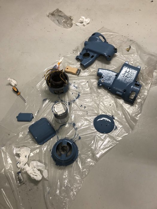 Delar av en demonterad pelarborr målade i blått spridda på en plastpresenning med målburkar och trasor.