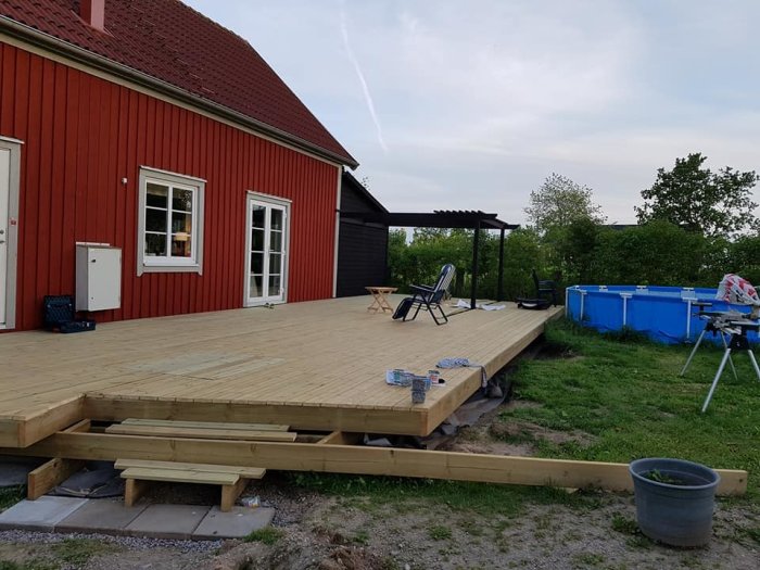 Nybyggd trädäck på 6 x 13 meter med pergola vid ett rött hus, tomten under uppbyggnad, byggmaterial synligt.