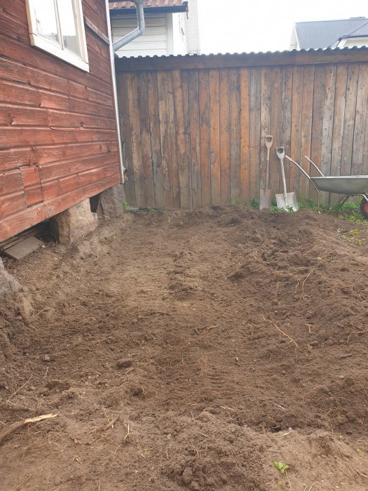 Uppgrävd mark för stenläggning nära ett rött skjul med spadar och en skottkärra.