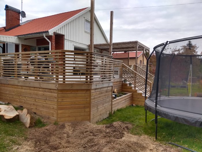 Nybyggd rymlig träaltan med räcke vid hus, flishög efter trädfräsning, trampolin till höger.