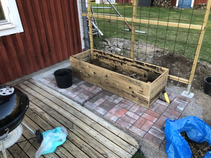 Nybyggd träodlingslåda på plattlagd mark med spaljé i bakgrunden vid rödfärgad husvägg.