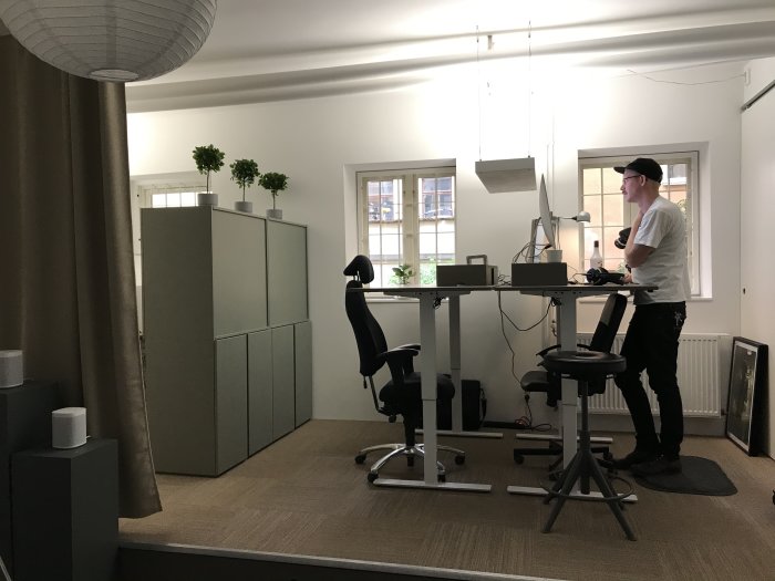 Interiör av ett kontor med stående person vid höj- och sänkbart skrivbord, datorskärmar och växter.