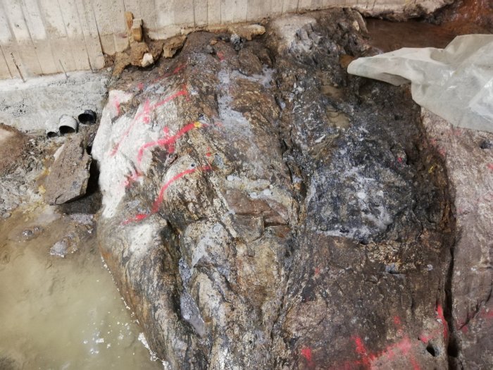 Stor sten med röda märkningar vid en byggarbetsplats, delvis omgiven av vatten och grus med rör synliga i bakgrunden.