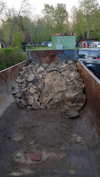 Lastbilskärra delvis fylld med stenar mot en bakgrund av träd och byggnader.
