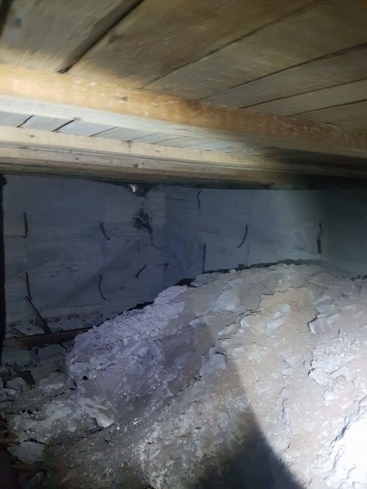Upplyst vy av ett dolt källarrum under trägolv, med berg och betongvägg som visar spår av vatteninträngning.