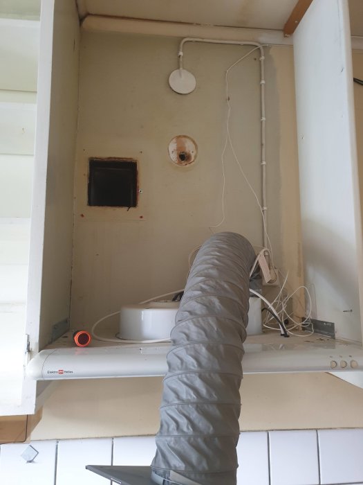 Öppet köksskåp med borttagen fläktkåpa, synlig icke-centrerad ventilation, elledningar och lägre tak bakom.