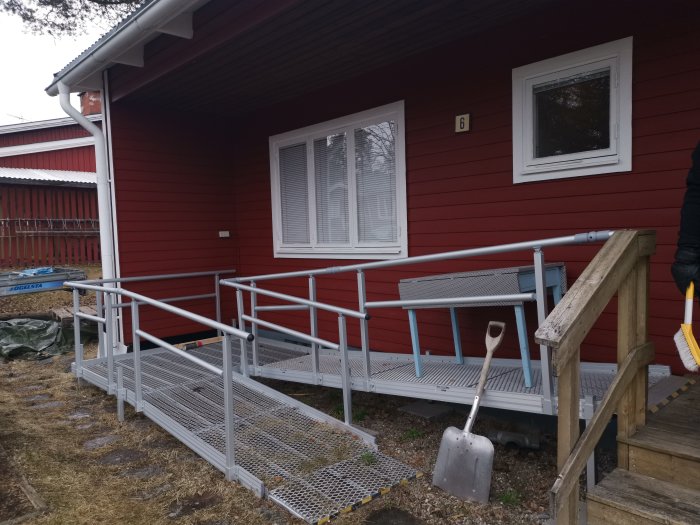 Metallramp avmonterad vid en rödmålad stuga, redo för försäljning, med spade i förgrunden.